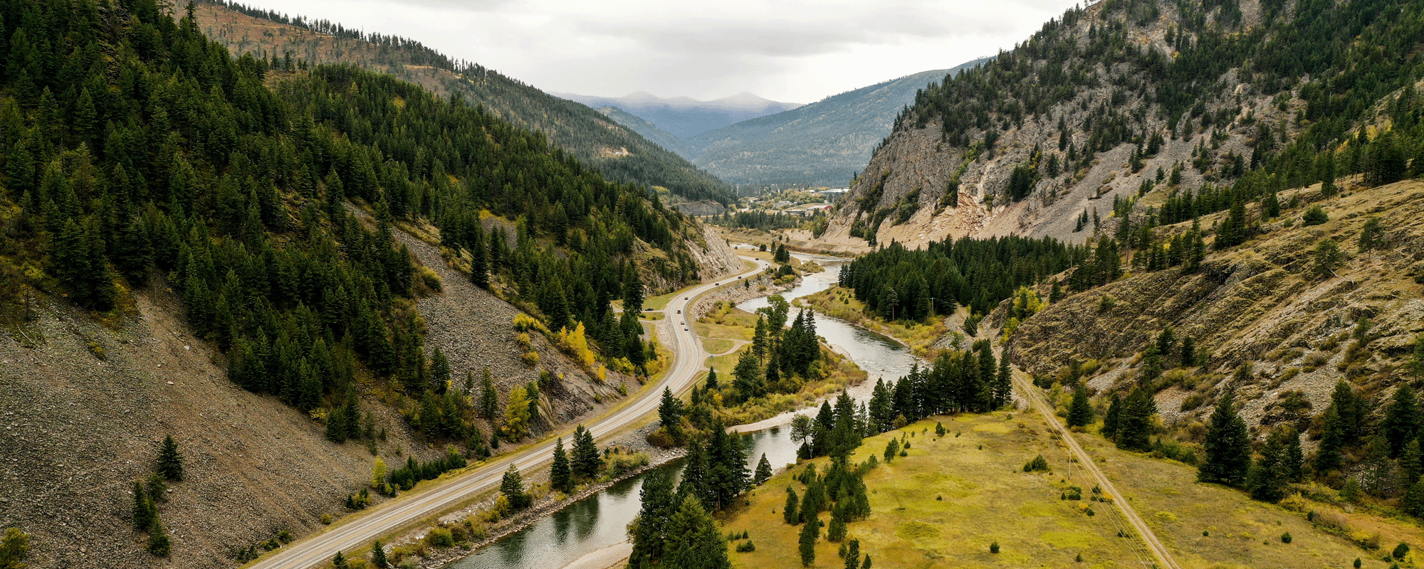 Attorneys. A scenic aerial view of a winding river and road passing through a lush, mountainous valley. The landscape is dotted with trees and patches of grassy meadows, with mountains visible in the background under a cloudy sky.