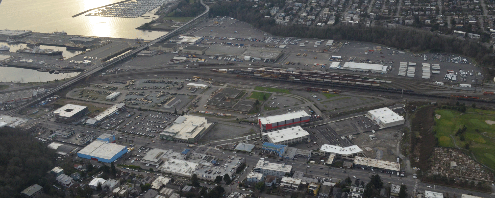 Community Planning & Policy Services. An aerial view of an industrial and residential area near a coastline, with a marina, railway yard, and buildings visible. The sun reflects off the water, and mountains are seen in the distance.