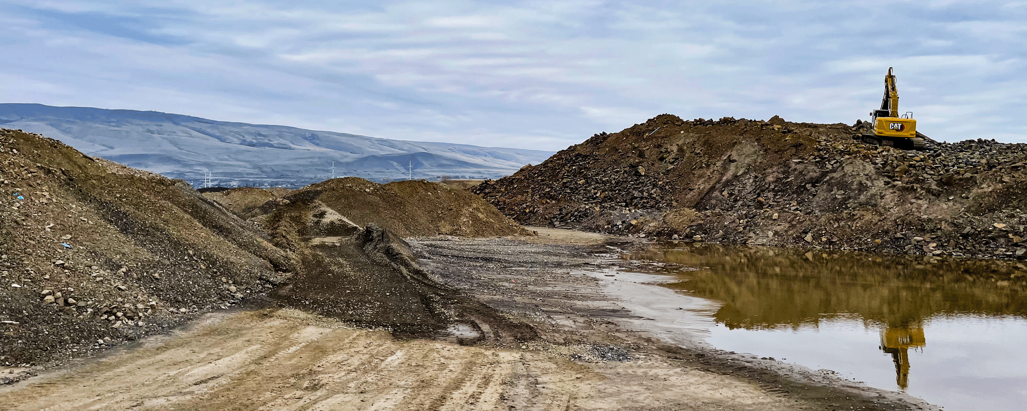 A construction site with dirt piles and a large excavator on a mound of earth. A muddy pathway and a water puddle are visible, with mountains in the background under a cloudy sky.