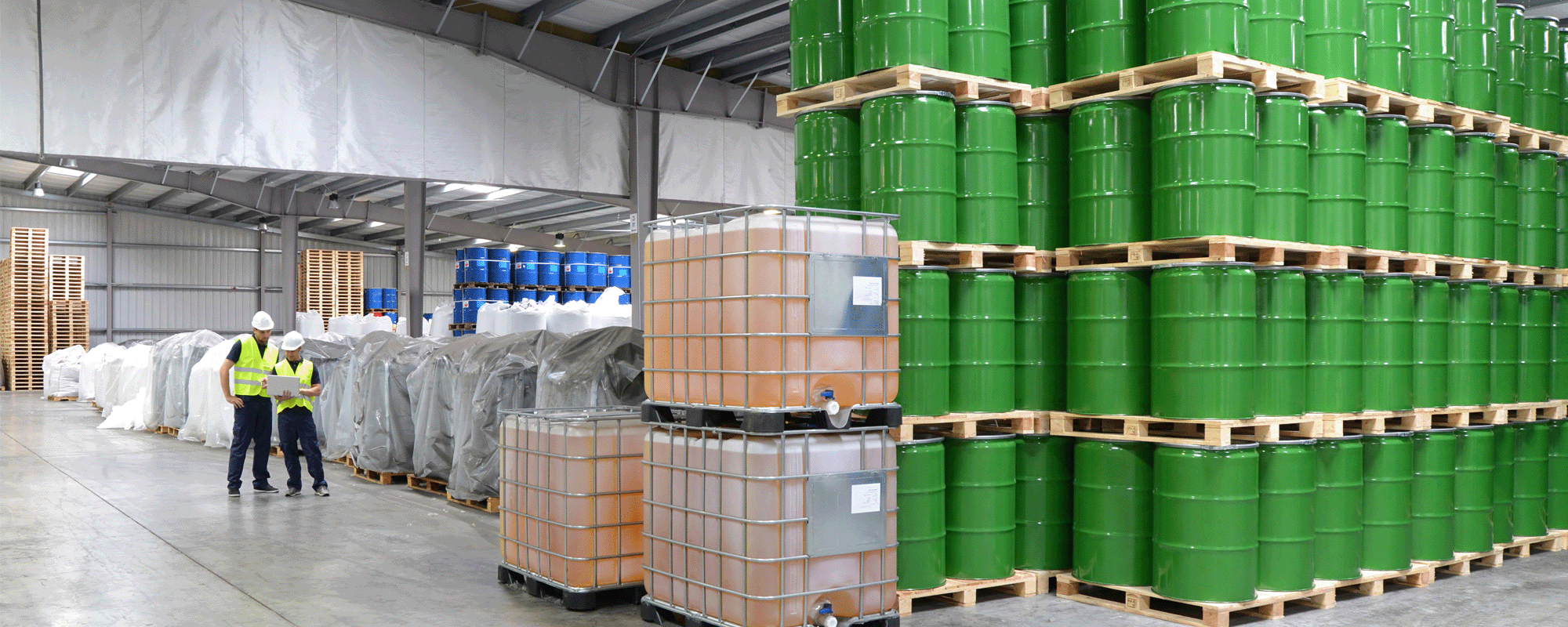 An industrial warehouse with stacks of green barrels and large containers. Two compliance workers in safety vests and helmets inspect a clipboard.