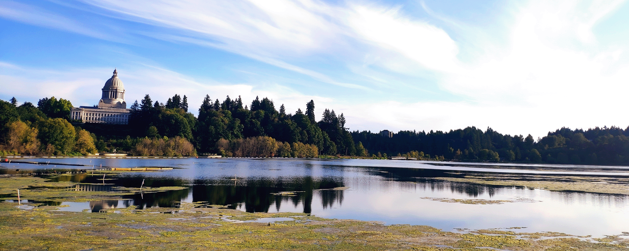  Local & State Agencies. A scenic view of a domed capitol building situated on a hill surrounded by trees, with a calm lake in the foreground reflecting the blue sky and wispy clouds.