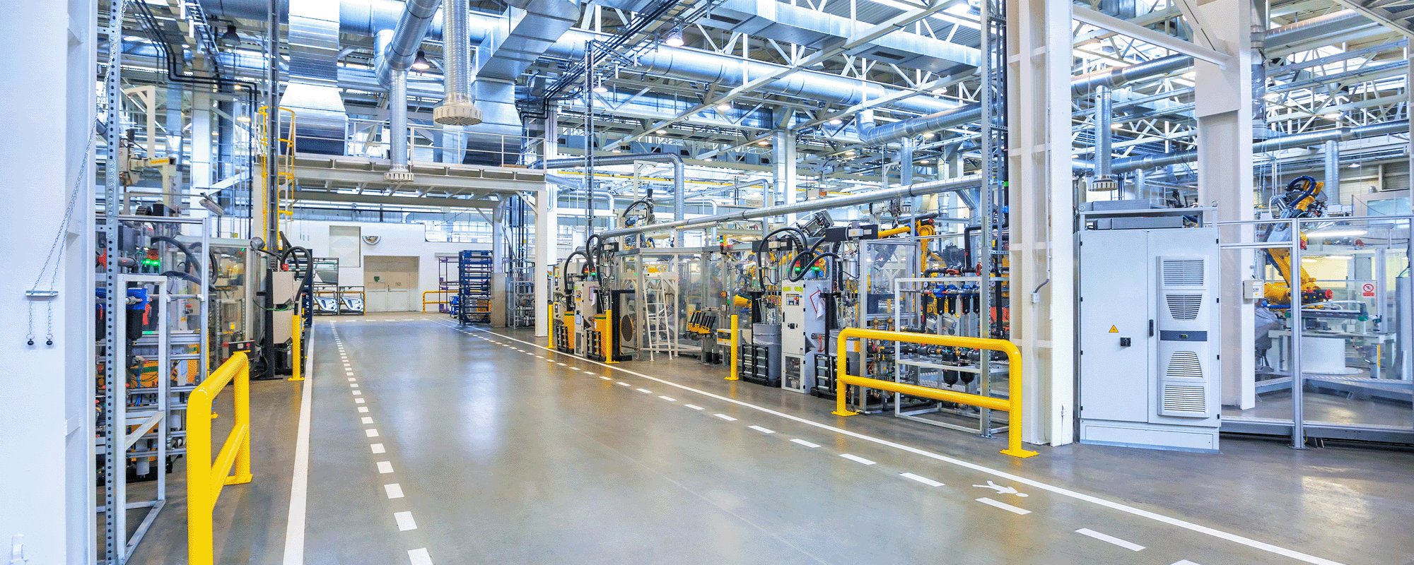 A brightly lit, modern manufacturing facility with high ceilings and extensive ductwork. Machinery and equipment are organized along the sides of a wide, marked walkway, which includes pedestrian symbols. The space is clean and well-maintained, with yellow safety railings and clear signage.