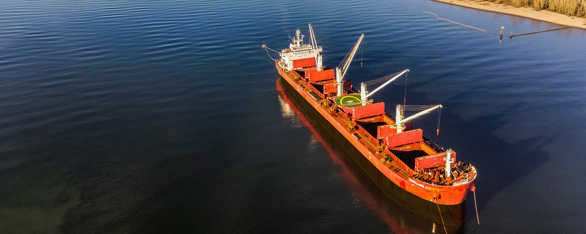 Ports. Aerial view of a large red cargo ship with cranes, sailing on a calm, wide river. The riverbanks are lined with fields and trees, with several other ships visible in the distance under a clear, blue sky.