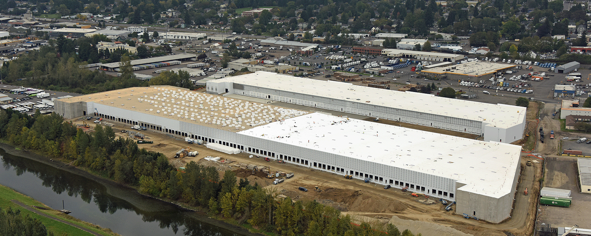 Real Estate Development. Aerial view of a large industrial warehouse complex under construction, with several white-roofed buildings. The site is bordered by a river and surrounded by a mix of industrial and residential areas, with a backdrop of rolling hills and a cloudy sky.