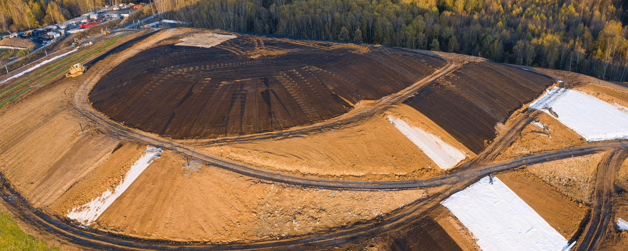 Aerial view of a solid waste and recycling site with sections of tilled soil, bare earth, and green grass. Curved dirt roads run through the site, surrounded by dense forest. Some areas are covered with white material.