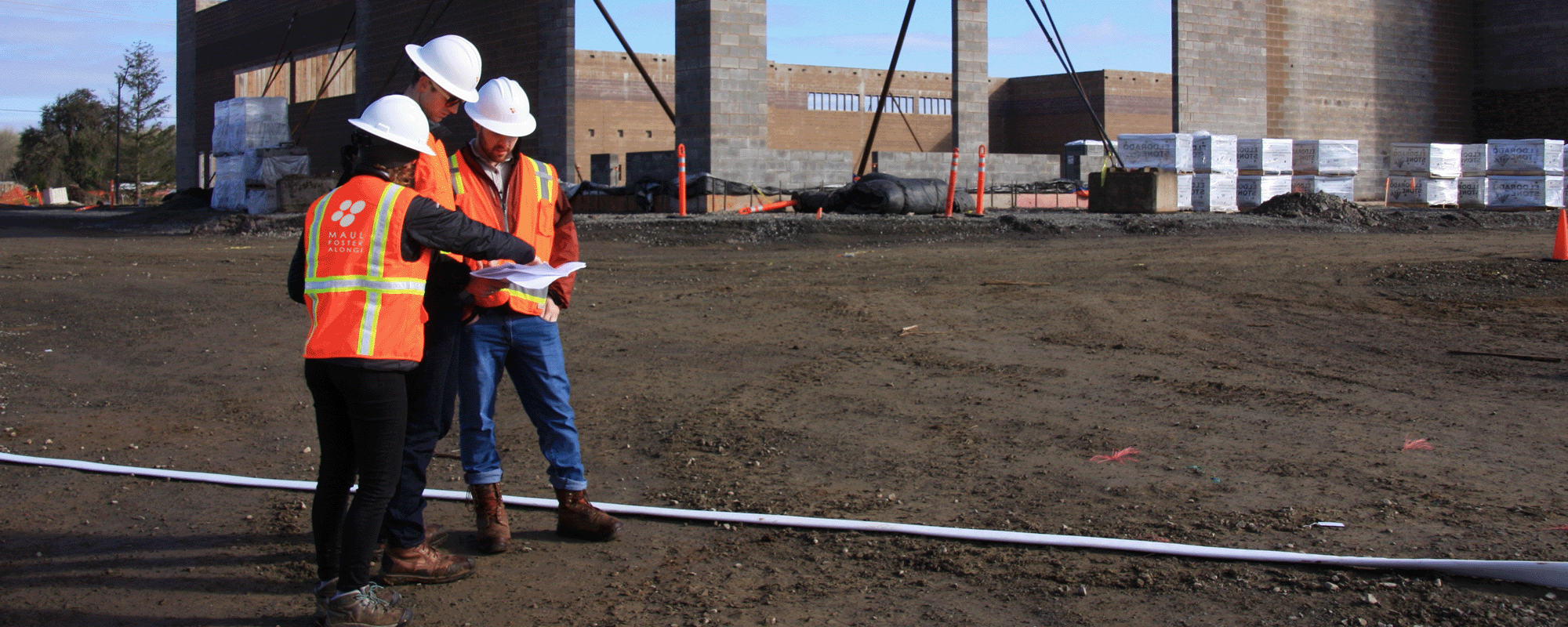 Three construction workers in safety vests and helmets review plans at a construction site with an unfinished building structure in the background.