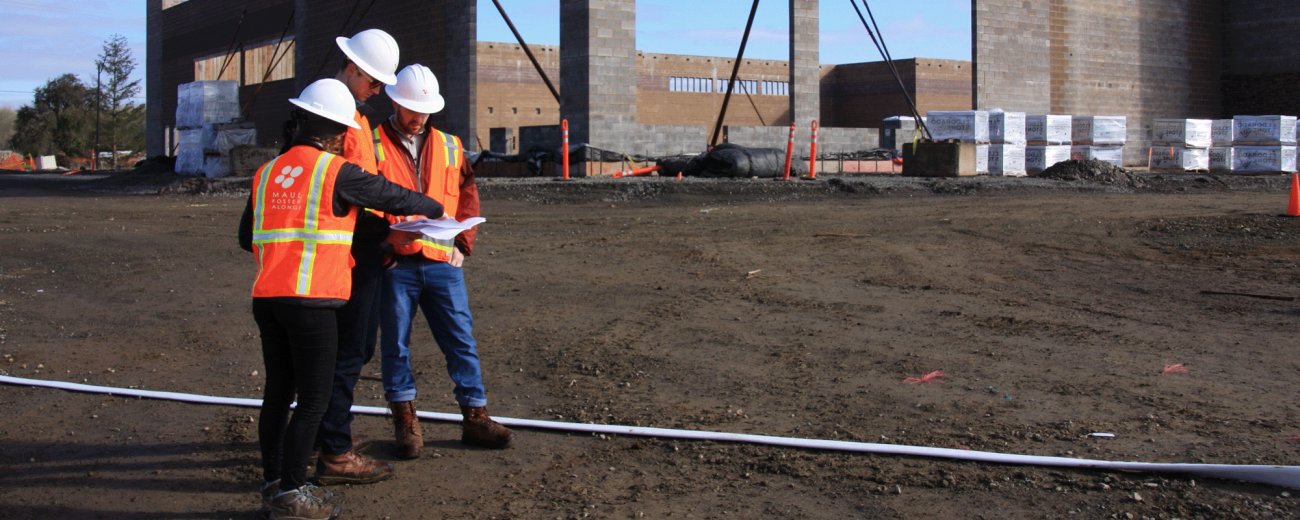 Three construction workers in safety vests and helmets review plans at a construction site with an unfinished building structure in the background.